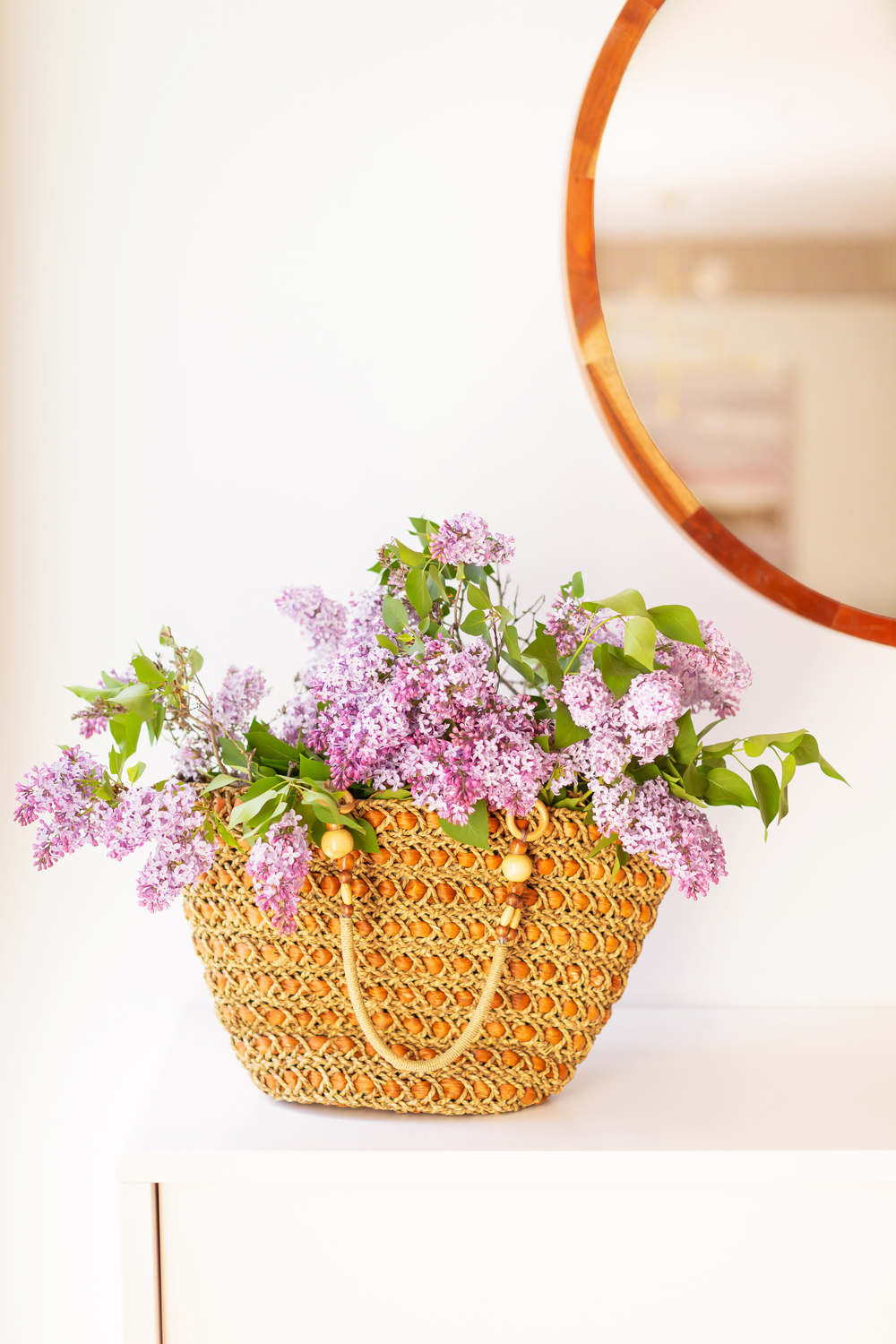 A vintage rattan tote bag overflowing with blooming lilac branches in JustineCelina’s light and airy mid century meets boho modern living room | DIY Lilac Flower Arrangement | How to Forage Lilacs | How to Prolong Lilac Vase Life | How to Arrange Lilacs | Fresh Lilac Flower Bouquet | Extremely Pretty Lilac Arrangement | Purple Lilac Arrangement | How to Cut Lilacs from a tree or bush | Calgary Creative Lifestyle Blogger // JustineCelina.com