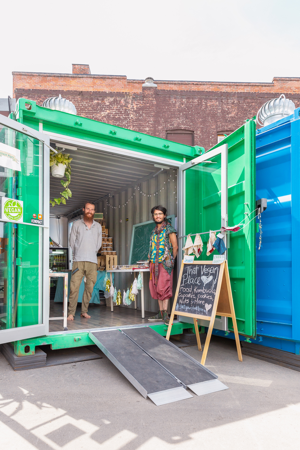 Exploring East Village Junction with Tourism Calgary | Vibrant Inglewood Calgary Summer Market with container stores, live performances, food trucks, vegan food, eclectic shopping, bike rentals and fitness classes | BARK Calgary Review | Two smiling men standing inside of a bright green shipping container converted into a vegan storefront at and outdoor market | Calgary Lifestyle Blogger // JustineCelina.com