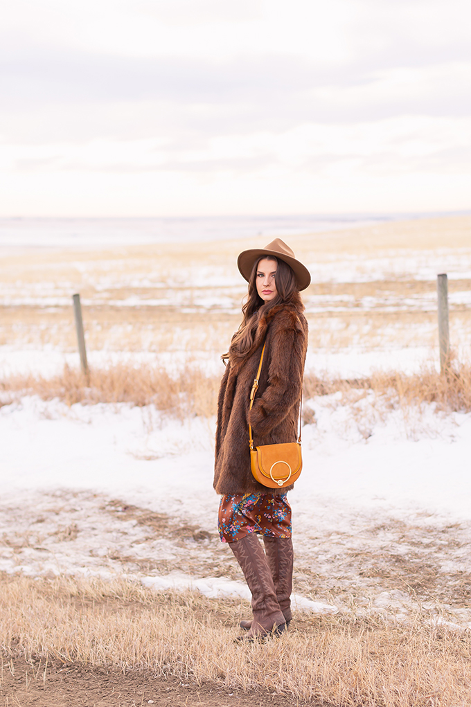 Winter 2019 Lookbook | Bohemian Rhapsody: How to Style Midi Dresses for Winter | Brunette Girl Standing in a Country Field at Sunrise Wearing a Brown Floral Dress, Faux Fur Coat, Brown Wide Brim Hat, Western OTK Boots and a Mustard Cross Body Bag | Bohemian Winter Style Ideas | Pantone Spring Summer 2019 Fashion Ideas // JustineCelina.com