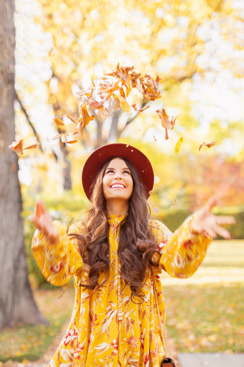 Early Autumn 2020 Lookbook |  Laughing brunette woman wearing a rust fedora and a mustard yellow botanical print shift dress throwing leaves into the air on a sunny autumn afternoon | warm weather fall outfit ideas | Fall 2020 Trend Guide | Casual Fall Fashion | Boho Fall 2020 Outfit Ideas | Fall in Calgary | Thanksgiving Outfit Ideas | | Transitional Summer Meets Autumn Fashion | October Style | Fall Fall Photoshoot | Calgary Alberta Fashion & Lifestyle Blogger // JustineCelina.com
