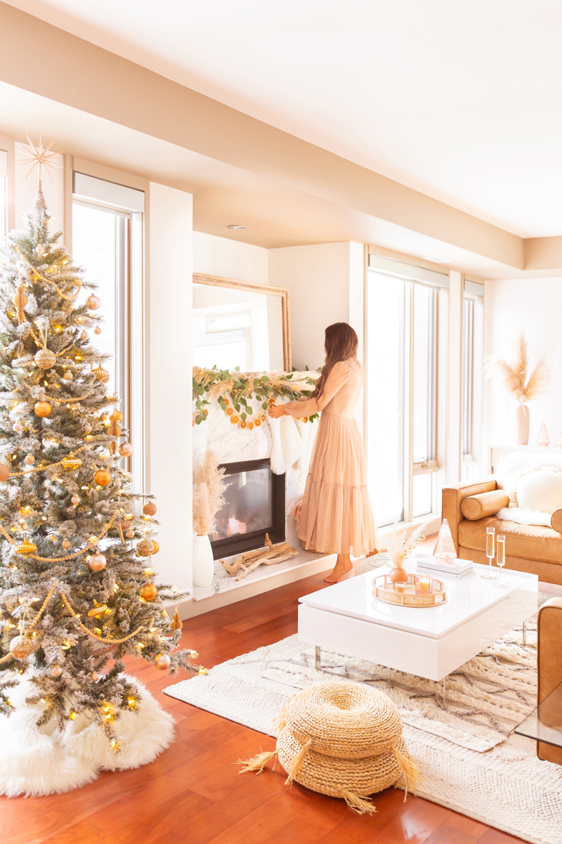 Mid Century Meets Boho Holiday Decor | Brunette woman wearing a taupe chiffon maxi dress hanging stockings in a bright and airy bohemian living room decorating for Christmas | Flocked Christmas Tree with Wood Garland, Metallic and Wood Ornaments | Bohemian Holiday Home Tour 2020 | Boho Chic Christmas Decor | DIY boho dried orange garland | Glam neutral holiday decor | Pampas Grass holiday decor | Pampas Grass Garland | Boho Christmas Tree | Canadian Tire CANVAS Ornaments // JustineCelina.com