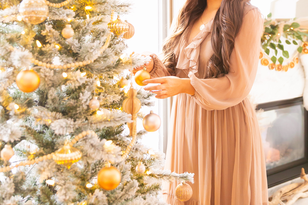 Mid Century Meets Boho Holiday Decor | Brunette woman wearing a taupe chiffon maxi dress decorating a flocked Christmas tree in an airy bohemian living room | Flocked Christmas Tree with Wood Garland, Metallic and Wood Ornaments | Bohemian Holiday Home Tour 2020 | Boho Chic Christmas Decor | DIY boho dried orange garland | Glam neutral holiday decor | Pampas Grass holiday decor | Pampas Grass Garland | Boho Christmas Tree // JustineCelina.com
