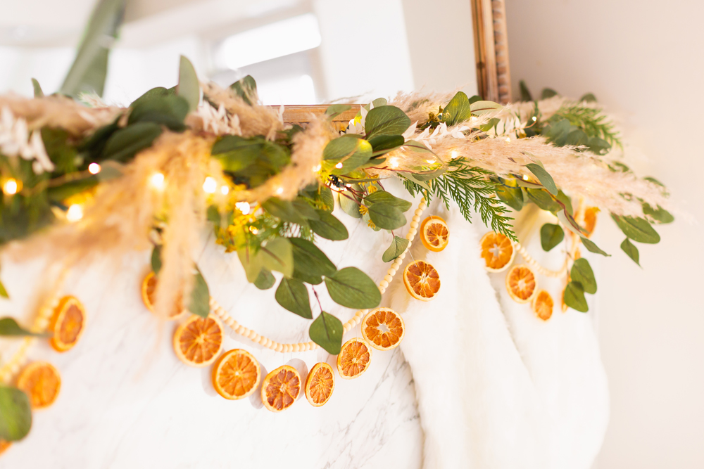 Mid Century Meets Boho Holiday Decor | Bright airy Mid Century Modern Living Room with a boho eucalyptus garland with pampas grass, fresh cedar, bleached Italian ruscus and a beaded dried orange garland | Boho Christmas Decorating Ideas for Apartments | Boho Holiday Fireplace Decor | DIY Orange Slice Garland | Bohemian Holiday Home Tour 2021 | Boho Chic Christmas Decor | Glam neutral holiday decor | Warm toned Holiday decor | Fun Holiday Decor | Dried Fruit Holiday Decor // JustineCelina.com