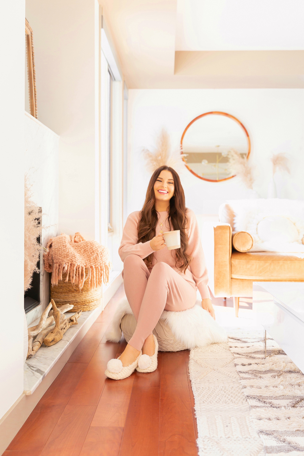 Smiling brunette woman wearing a blush pink lounge set and cream slipper sitting on a velvet pouf with cup of tea in a bright and airy boho living room | Calgary Lifestyle Blogger // JustineCelina.com
