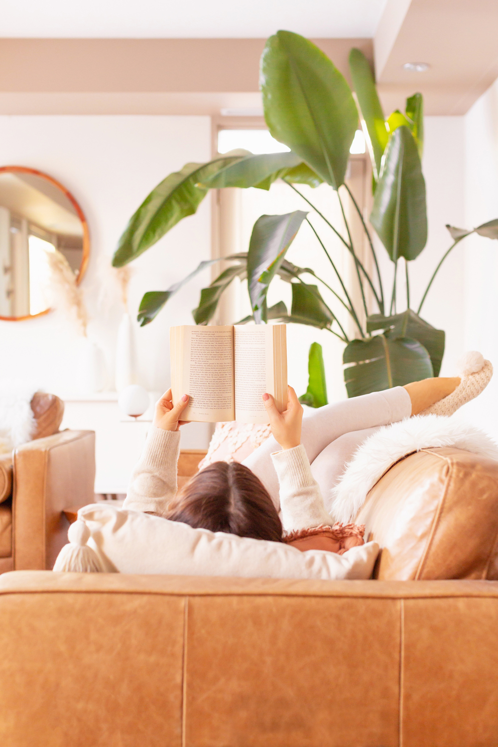 Brunette woman in cream loungewear laying on a caramel leather couch reading a book in a bright and airy boho living room with a mature Bird of Paradise Plant in the background | Best Self Help Books | Best Audible Books | Motivational Books | Inspirational Books | Inspirational Books for Women | Books for Female Entrepreneurs | Motivational Books for Creatives | Best Self Help Books for Women | Best Personal Growth Books | Self Healing Books | Calgary Lifestyle Blogger // JustineCelina.com