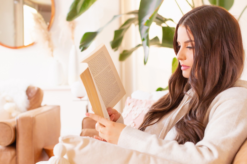 Brunette woman in cream loungewear laying on a caramel leather couch reading a book in a bright and airy boho living room with a mature Bird of Paradise Plant in the background | Best Self Help Books | Best Audible Books | Motivational Books | Inspirational Books | Inspirational Books for Women | Books for Female Entrepreneurs | Motivational Books for Creatives | Best Self Help Books for Women | Best Personal Growth Books | Self Healing Books | Calgary Lifestyle Blogger // JustineCelina.com