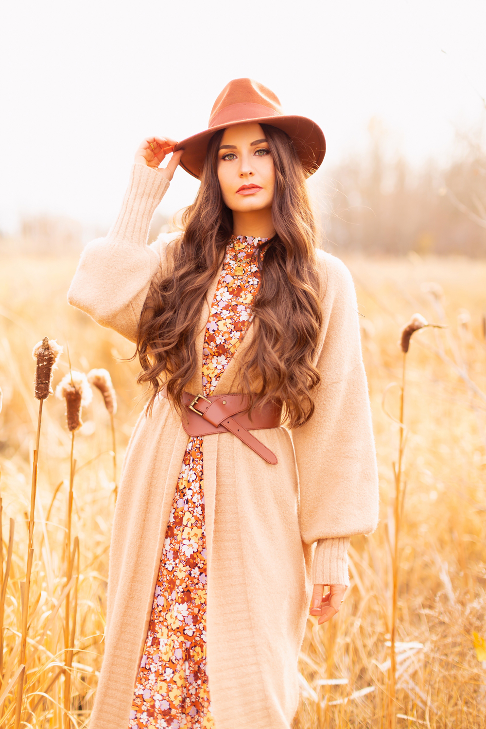 6 Luxe Bohemian Fall Outfits | Elegant Equestrian | Brunette woman wearing a fall floral midi dress, a longline camel cardigan, a cognac leather corset belt, cognac western knee high boots, a rust fedora and a round cognac crossbody bag amongst golden grasses and cattails | Boho Fall 2022 Outfit Ideas | Classic Fall Outfit Formulas | Feminine Bohemian Style | Polished Bohemian Style | Elegant Bohemian Style | Bohemian Outfit Ideas | Calgary Alberta Fashion Blogger // JustineCelina.com