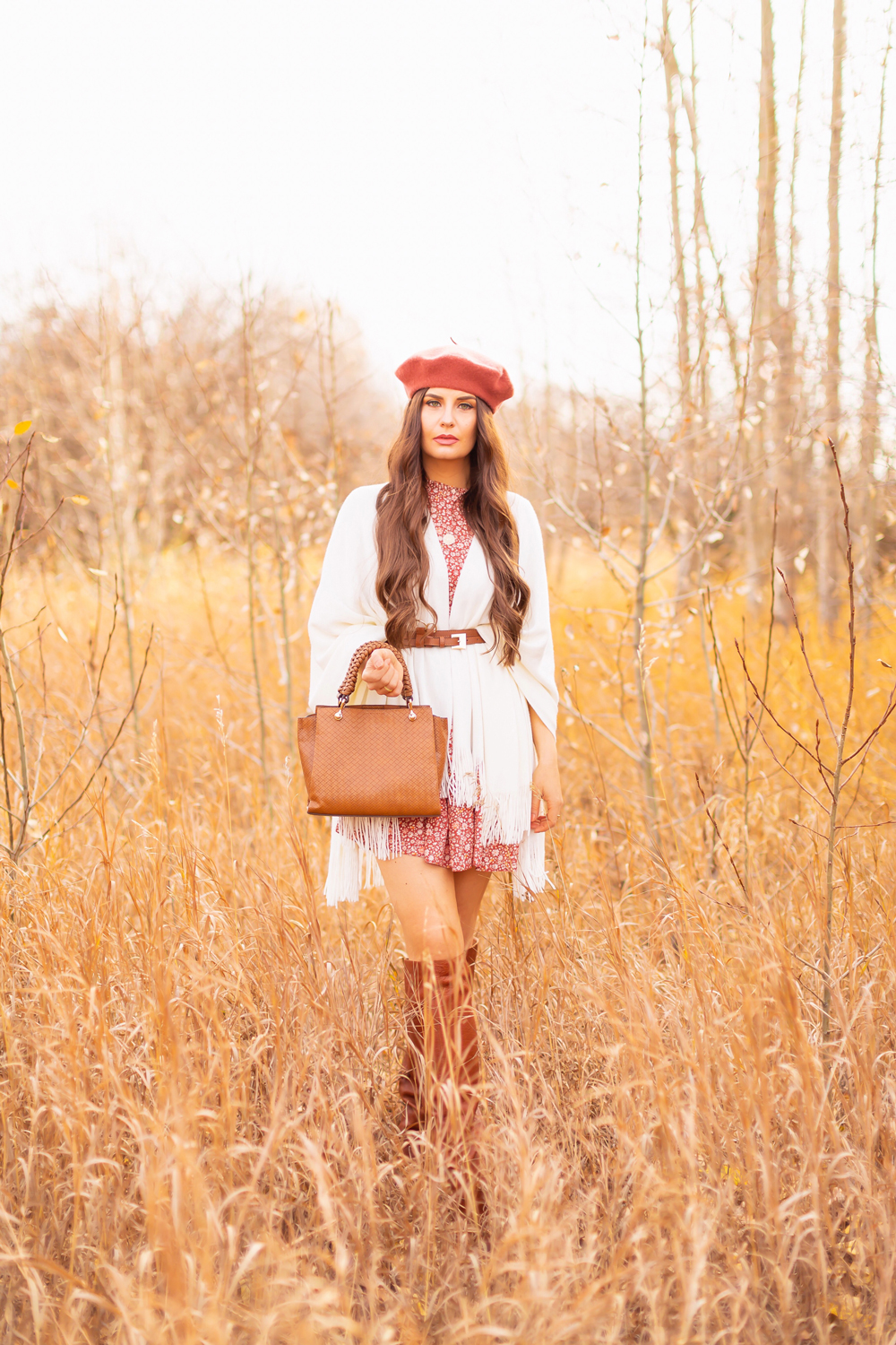 6 Luxe Bohemian Fall Outfits | Playful Poncho | Brunette woman wearing a fall floral mini dress, a cream fringe trimmed poncho, a cognac leather corset belt, cognac western knee high boots, a rust fedora and a woven cognac crossbody bag amongst golden grasses and trees | Boho Fall 2022 Outfit Ideas | Classic Fall Outfit Formulas | Feminine Bohemian Style | Polished Bohemian Style | Elegant Bohemian Style | Bohemian Outfit Ideas | Calgary Alberta Fashion Blogger // JustineCelina.com