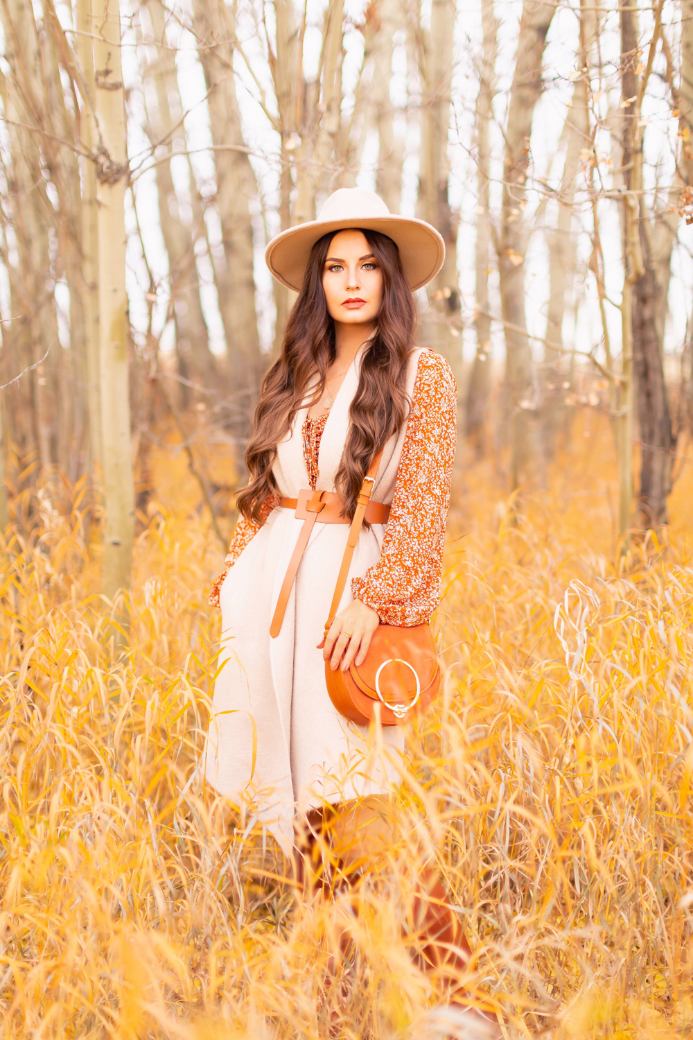6 Luxe Bohemian Fall Outfits | Rancher Revival | Brunette woman wearing an orange floral mini dress, beige Pink Martini Stockport Vest, wide cognac waist belt with tie detail, a crossbody cognac suede saddle bag and Lucky Brand Whiskey Azoola Leather Boot amoungst golden grasses and bare trees | Boho Fall 2022 Outfit Ideas | Classic Fall Outfit Formulas | Feminine Bohemian Style | Bohemian Outfit Ideas | How to Style a Rancher Hat | Calgary Alberta Fashion Blogger // JustineCelina.com