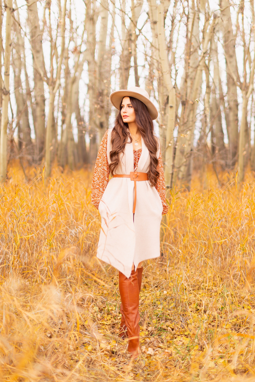 6 Luxe Bohemian Fall Outfits | Rancher Revival | Brunette woman wearing an orange floral mini dress, beige Pink Martini Stockport Vest, wide cognac waist belt with tie detail, a crossbody cognac suede saddle bag and Lucky Brand Whiskey Azoola Leather Boot amoungst golden grasses and bare trees | Boho Fall 2022 Outfit Ideas | Classic Fall Outfit Formulas | Feminine Bohemian Style | Bohemian Outfit Ideas | How to Style a Rancher Hat | Calgary Alberta Fashion Blogger // JustineCelina.com