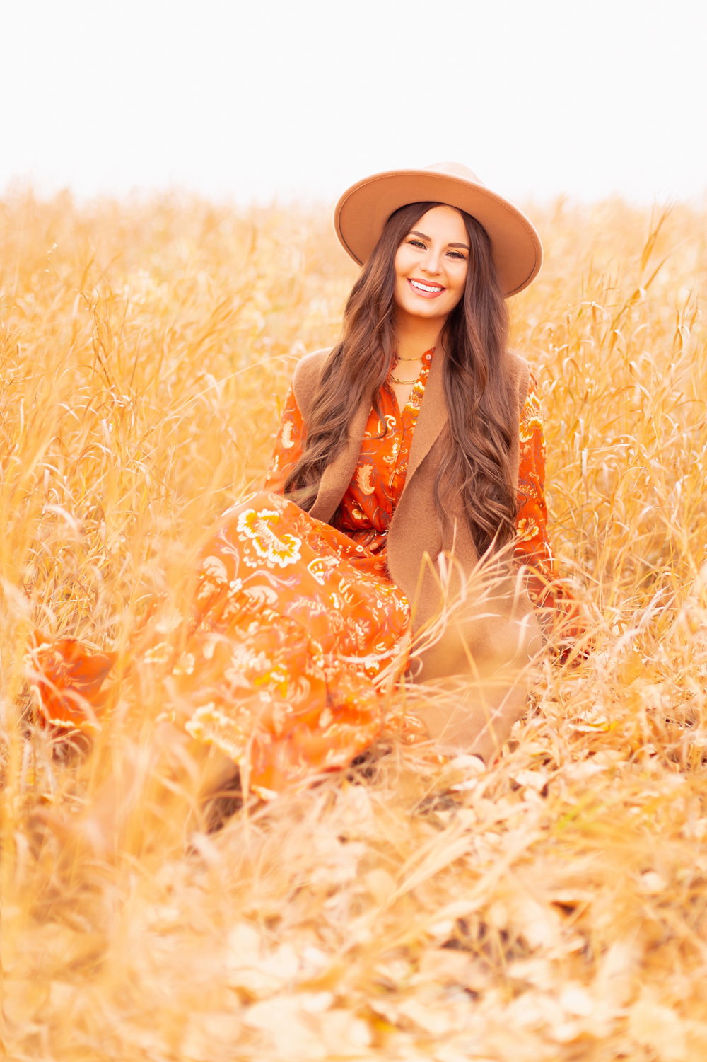 6 Luxe Bohemian Fall Outfits | Thanksgiving Chic | Smiling brunette woman sitting in golden wild grasses wearing an orange paisley midi dress, brown Pink Martini Stockport Vest and a camel flat brimmed rancher hat | Boho Fall 2022 Outfit Ideas | Classic Fall Outfit Formulas | Feminine Bohemian Style | Polished Bohemian Style | Elegant Bohemian Style | Bohemian Outfit Ideas | Calgary Alberta Fashion Blogger // JustineCelina.com