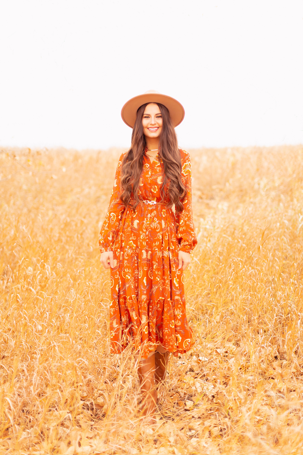 6 Luxe Bohemian Fall Outfits | Thanksgiving Chic | Smiling brunette woman wearing an orange paisley midi dress, brown Pink Martini Stockport Vest, a slim cognac leather corset, a vintage leather hand tooled bag and Vince Camuto Cognac Senimda Mid Calf Boot amoungst golden grasses | Boho Fall 2022 Outfit Ideas | Classic Fall Outfit Formulas | Feminine Bohemian Style | Polished Bohemian Style | Elegant Bohemian Style | Bohemian Outfit Ideas | Calgary Alberta Fashion Blogger // JustineCelina.com