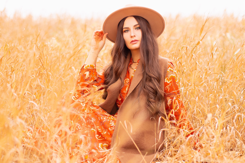 6 Luxe Bohemian Fall Outfits | Thanksgiving Chic | Smiling brunette woman sitting in golden wild grasses wearing an orange paisley midi dress, brown Pink Martini Stockport Vest and a camel flat brimmed rancher hat | Boho Fall 2022 Outfit Ideas | Classic Fall Outfit Formulas | Feminine Bohemian Style | Polished Bohemian Style | Elegant Bohemian Style | Bohemian Outfit Ideas | Calgary Alberta Fashion Blogger // JustineCelina.com